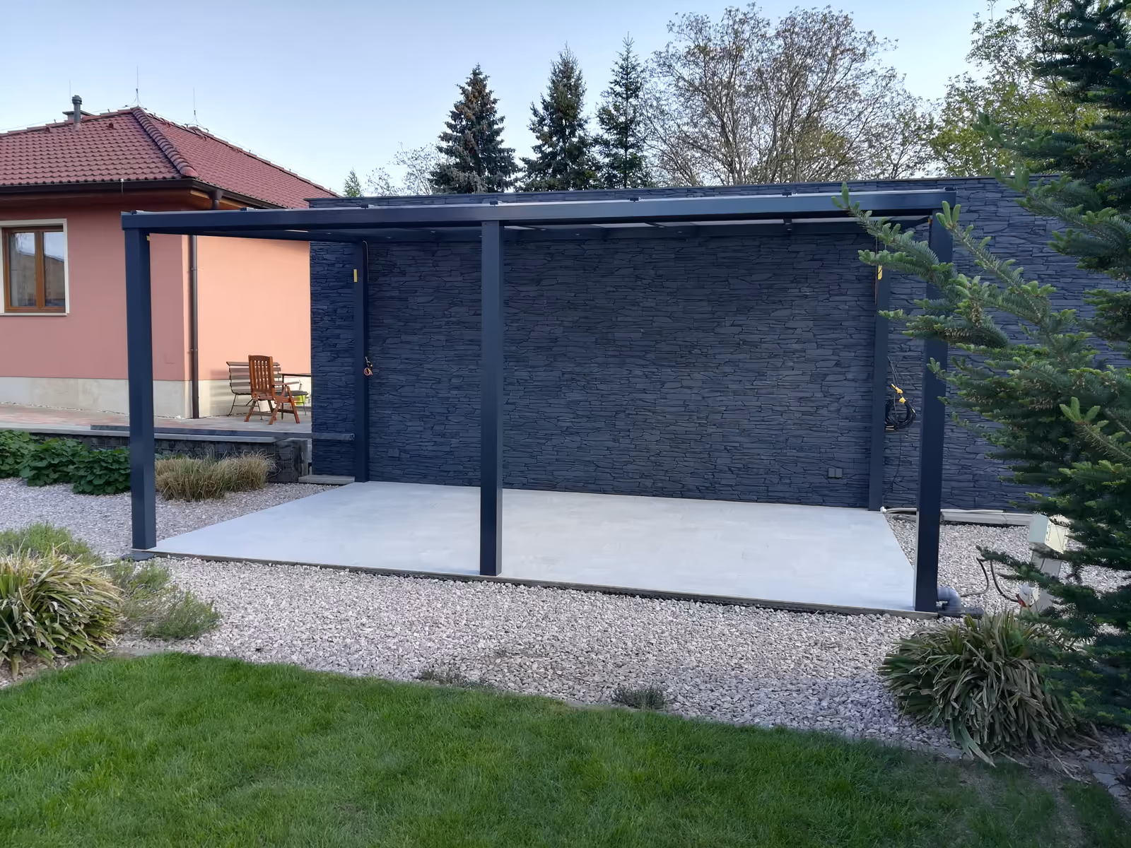 antracitová pergola nad šedou dlažbou před kamennou stěnou
